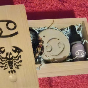 May include: A wooden box with a Cancer zodiac symbol on the front. The box is open and contains a small, round, beige soap with a Cancer zodiac symbol, a small bottle of essential oil with a Cancer zodiac symbol, and a gold-coloured charm with a Cancer zodiac symbol.
