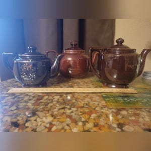 Può includere: Tre teiere in ceramica in varie tonalità di marrone. La teiera a sinistra è blu scuro con un motivo floreale. La teiera centrale è marrone medio e la teiera a destra è marrone più scuro. Un righello di legno è di fronte alle teiere.
