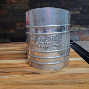 Może przedstawiać: Zabytkowe, cylindryczne metalowe sito z wytłoczonym napisem "SIFT-CHINE" i "GUARANTEED SATISFACTORY MADE IN U.S.A." z boku. Sito jest srebrne i ma poziome paski.