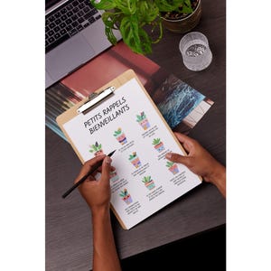 May include: A wooden clipboard with a printed sheet featuring illustrated potted plants and the text "PETITS RAPPELS BIENVEILLANTS". A person is holding the clipboard and writing with a black pen. A laptop and a potted plant are in the background.