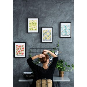 May include: Four framed prints with floral patterns hang on a gray wall. The prints feature different floral designs in shades of yellow, red, white, and blue. The prints are arranged in a grid pattern. A person is sitting in front of the prints, facing away from the camera.