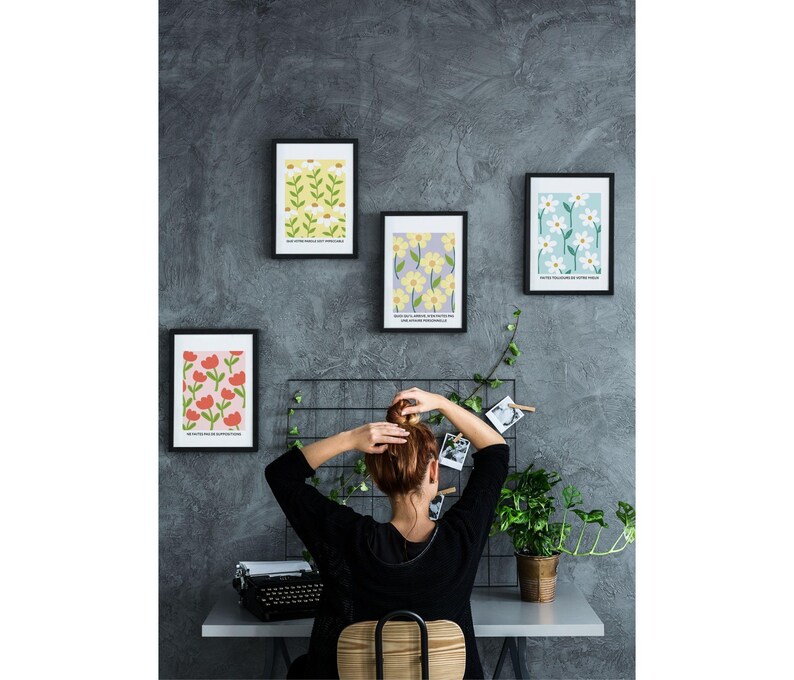 May include: Four framed prints with floral patterns hang on a gray wall. The prints feature different floral designs in shades of yellow, red, white, and blue. The prints are arranged in a grid pattern. A person is sitting in front of the prints, facing away from the camera.