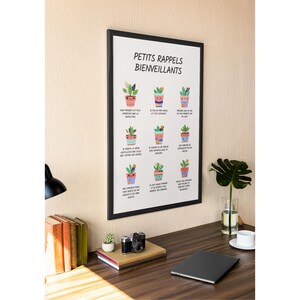 May include: A framed print with the text "PETITS RAPPELS BIENVEILLANTS" and illustrations of potted plants. The print is displayed on a wooden desk with a lamp, books, a camera, and a laptop. The wall is a light beige color.