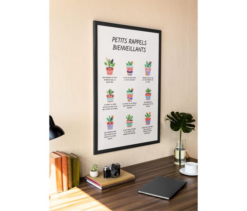 May include: A framed print with the text "PETITS RAPPELS BIENVEILLANTS" and illustrations of potted plants. The print is displayed on a wooden desk with a lamp, books, a camera, and a laptop. The wall is a light beige color.