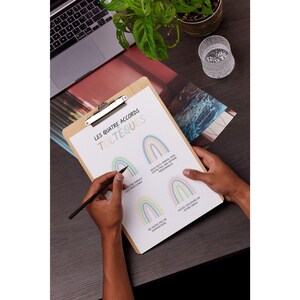 May include: A clipboard with a white sheet of paper on it. The paper has the title "Les Quatre Accords Tolteques" written in black. There are four rainbow illustrations on the paper, each with a different colored background. The text below each rainbow is in French.