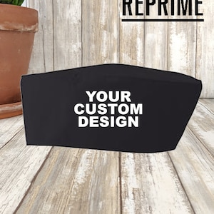 May include: Black face mask with the text "YOUR CUSTOM DESIGN" in white. The mask is displayed on a wooden surface. The Artisan Collection by Reprime logo is visible in the background. A potted plant is in the background.