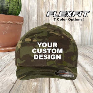 Peut inclure: Un chapeau Flexfit camouflage vert avec le texte "YOUR CUSTOM DESIGN" en blanc. Le chapeau a un logo Flexfit noir au dos.
