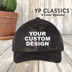 May include: A black and green camouflage baseball cap with a black mesh back. The cap has white text that says "YOUR CUSTOM DESIGN".