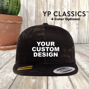May include: Black and green camouflage baseball cap with a black brim and a yellow stripe. The cap has the text "YOUR CUSTOM DESIGN" in white letters on the front. The cap is made by "The Classic Snapback".