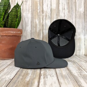 May include: A dark grey baseball cap with a small triangle logo on the front. The cap is shown from the side and the back of the cap is visible.