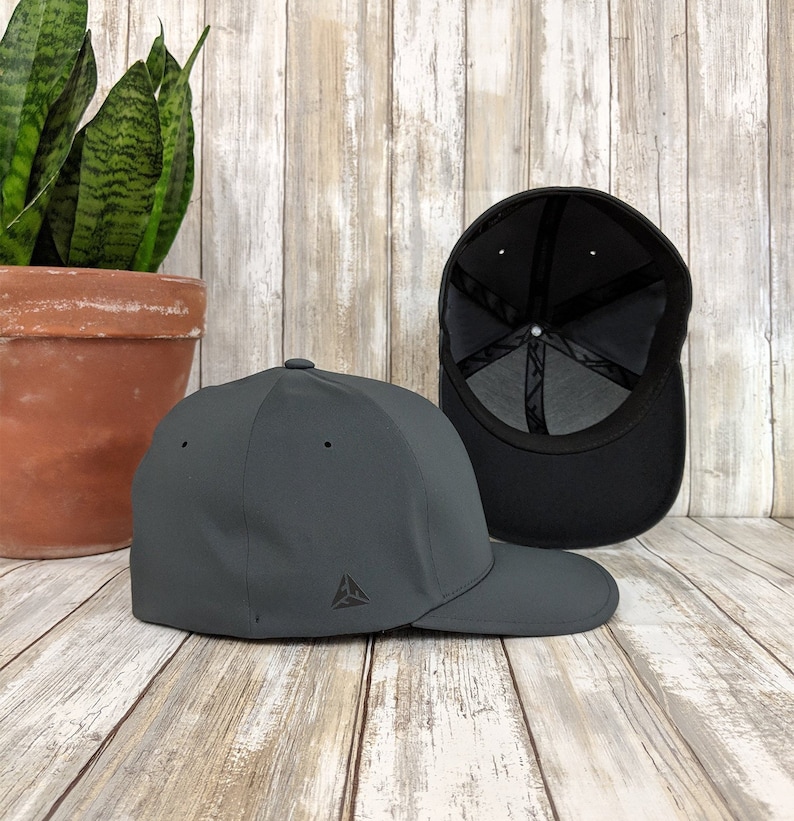 May include: A dark grey baseball cap with a small triangle logo on the front. The cap is shown from the side and the back of the cap is visible.
