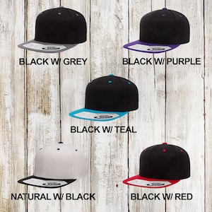 May include: Five different colour variations of a black and white baseball cap. The caps are all flat-brimmed and have a contrasting colour under the brim. The colours are black with grey, black with purple, black with teal, natural with black, and black with red.