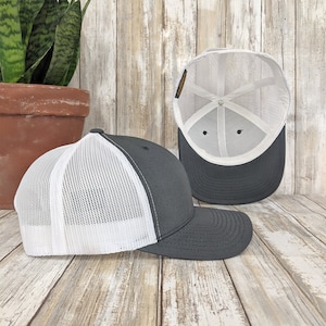 May include: A gray and white trucker hat with a mesh back. The hat is shown from two angles, one with the front of the hat facing the camera and the other with the inside of the hat facing the camera.
