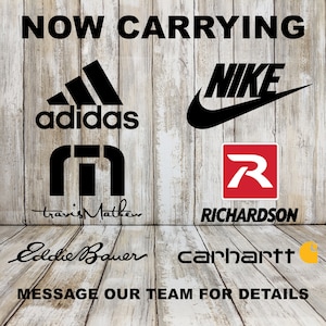 Op de afbeelding: Een houten achtergrond met de tekst "NOW CARRYING" in zwarte letters. Onder de tekst staan logo's van Adidas, Nike, Travis Mathew, Richardson, Eddie Bauer en Carhartt.