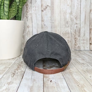 Op de afbeelding: Een zwarte baseballpet met een bruin leren bandje. Het bandje heeft het woord "REVERSE" erop gegraveerd.