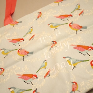 Peut inclure: Un sac fourre-tout bleu clair avec un motif répété d'oiseaux colorés et d'éléments floraux. Le sac a des poignées corail et le texte "It's a lovely Day" imprimé sur le devant.