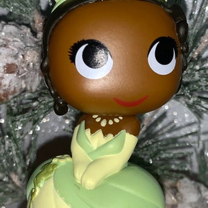 May include: A green and yellow Tiana princess ornament with a brown face and large eyes. The ornament is a stylized version of the Disney princess.