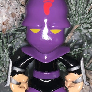 May include: Purple and black Christmas ornament of a ninja figure with a red footprint on the head. The figure has yellow eyes and is holding a silver weapon.