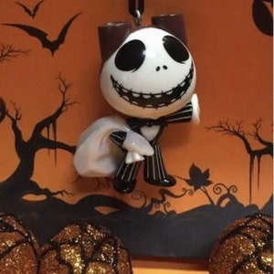 May include: A white and black ceramic ornament of Jack Skellington from the Nightmare Before Christmas movie. He is holding a white bag in his left hand and his right hand is raised. He is wearing a black and white striped suit and a black bow tie.