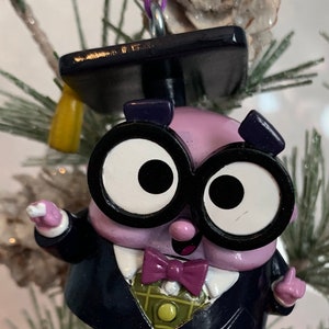 May include: Purple character wearing a black graduation cap and gown, black framed glasses, and a purple bow tie. The character has large white eyes and a big smile.