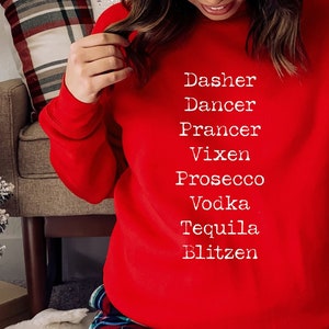 May include: A red sweatshirt with white text that reads "Dasher, Dancer, Prancer, Vixen, Prosecco, Vodka, Tequila, Blitzen". The person is wearing a white knit beanie with a brown pom pom and red and black plaid pyjama trousers.