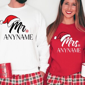 May include: A white and red Christmas themed sweatshirt with the text "Mr. ANYNAME" and a red Santa hat. A red sweatshirt with the text "Mrs. ANYNAME" and a red Santa hat. Both sweatshirts have a snowflake design.