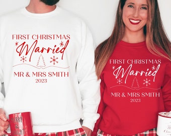 May include: A couple wearing matching white and red Christmas jumpers with the text "First Christmas Married" and their last name and the year 2023. The jumpers have a small Christmas tree and snowflakes.