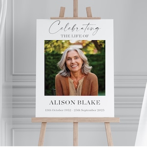 May include: White memorial sign with a black border, celebrating the life of Alison Blake. The sign includes a photo of Alison, the dates 13th October 1952 - 25th September 2025, and the text "Celebrating the life of".