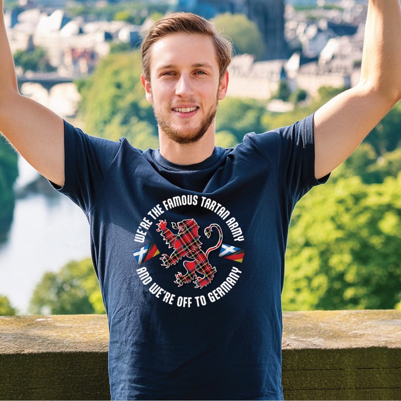 Puede incluir: Una camiseta azul marino con un gr&aacute;fico circular blanco que presenta un le&oacute;n de tart&aacute;n rojo y verde, dos banderas y el texto "We're the Famous Tartan Army and We're Off to Germany".