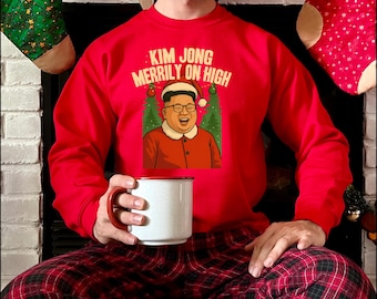 Puede incluir: Una sudadera roja con el texto "KIM JONG MERILLY ON HIGH" y una imagen de dibujos animados de Kim Jong-un con un gorro de Papá Noel. La persona de la imagen sostiene una taza blanca con borde y asa rojos. La persona lleva un pantalón de pijama a cuadros rojos y negros.
