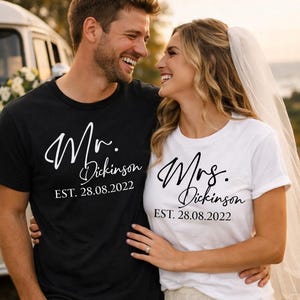 Puede incluir: Una pareja sonriente con camisetas de boda a juego. La camiseta negra dice "Mr. Dickinson EST. 28.08.2022" en escritura blanca. La camiseta blanca dice "Mrs. Dickinson EST. 28.08.2022" en escritura negra. Ropa de boda.