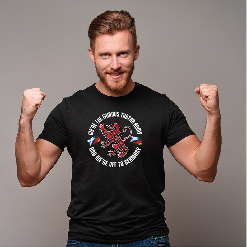 Puede incluir: Una camiseta negra con un gr&aacute;fico circular blanco que presenta un le&oacute;n de tart&aacute;n rojo y negro con el texto "We're the Famous Tartan Army and We're Off to Germany".