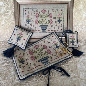 May include: A framed cross-stitch sampler with pink roses, blue vases, and an alphabet. Accompanying items include a small pillow, a rectangular piece, and a tag, all with matching floral designs and navy tassels. Gold scissors are also present.