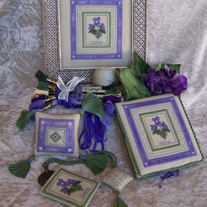 May include: A set of four needlepoint projects featuring a purple violet flower design. The projects include a framed piece, a small pillow, a small book, and a small pouch. The projects are all in shades of purple, green, and white.