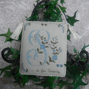 May include: A white cross-stitch sampler with a blue and green embroidered letter 'S' and the text 'S is for Sincerity'. The sampler is hanging from a black metal hook with green ivy vines.