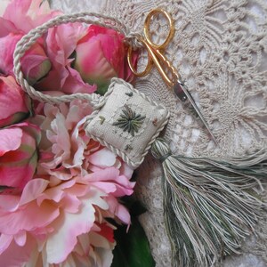 Puede incluir: Una pequeña almohada blanca bordada con un diseño floral verde y marrón, unida a una larga borla verde y blanca. La almohada está sobre un encaje blanco de ganchillo con flores rosas.