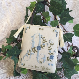 以下が含まれることがあります： 青い花と緑の花で囲まれたアルファベットの「A」が刺繍された白いクロスステッチのサンプル。文字の下には「A is for Affection」と刺繍されています。