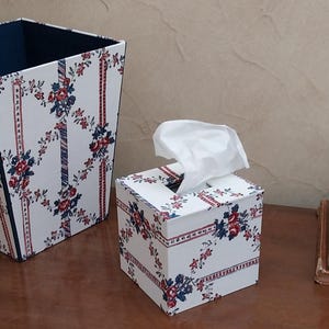 May include: A square tissue box cover and a matching tall wastebasket, both featuring a white background with a repeating pattern of red and blue floral sprigs and vertical striped ribbons. The tissue box has a white tissue protruding from the top.