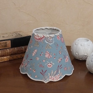 Peut inclure: Abat-jour bleu avec un motif floral rose et blanc. Le bord de l'abat-jour est festonné et il possède une armature métallique. Il est posé sur une surface en bois.
