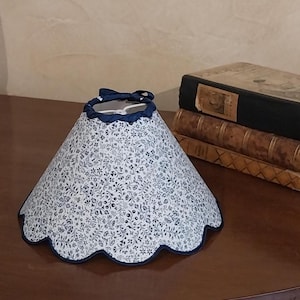 Peut inclure: Un abat-jour conique avec un bord festonné. Il présente un fond blanc orné d'un motif floral bleu. La bordure et le bord supérieur sont bleu marine. Un petit nœud bleu marine orne le sommet.