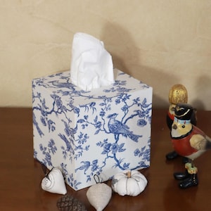Pode incluir: Caixa de lenços quadrada com um padrão floral e de pássaros em azul e branco. Os lenços brancos são visíveis no topo. Itens decorativos, incluindo uma pequena abóbora e estatuetas de pássaros, são dispostos ao redor.