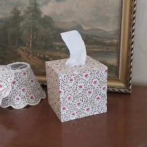 May include: A square tissue box cover featuring a repeating pattern of small pink flowers and dark green leaves on a cream background. A single white tissue protrudes from the top opening. Beside it sits a matching scalloped fabric lampshade.