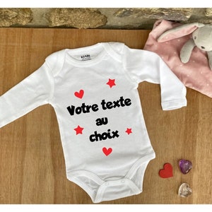May include: White long-sleeve baby onesie with red hearts and stars. The onesie has black text that says "Votre texte au choix".