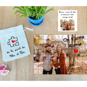 May include: A personalized jigsaw puzzle with a photo of a family of four, including two young children. The puzzle is in a white bag with a red heart and the text "de la part de El&eacute;a et L&eacute;o". The puzzle also has the text "On t'aime Mamie" with a red heart.