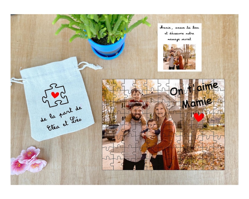 May include: A personalized jigsaw puzzle with a photo of a family of four, including two young children. The puzzle is in a white bag with a red heart and the text "de la part de El&eacute;a et L&eacute;o". The puzzle also has the text "On t'aime Mamie" with a red heart.
