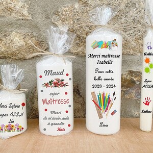 May include: Four white candles with personalized messages for teachers. The candles feature colorful illustrations and text such as "Merci Maitresse", "Super AESH", "Maud", and "Ashley".