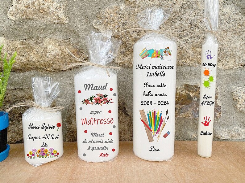 May include: Four white candles with personalized messages for teachers. The candles feature colorful illustrations and text such as "Merci Maitresse", "Super AESH", "Maud", and "Ashley".