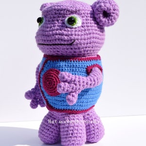May include: Purple crocheted stuffed toy with blue and burgundy accents. The toy has large green eyes and a small smile. The toy is standing on a white surface. The text "THECROCHETBOXSTUDIO" is visible on the toy's chest.