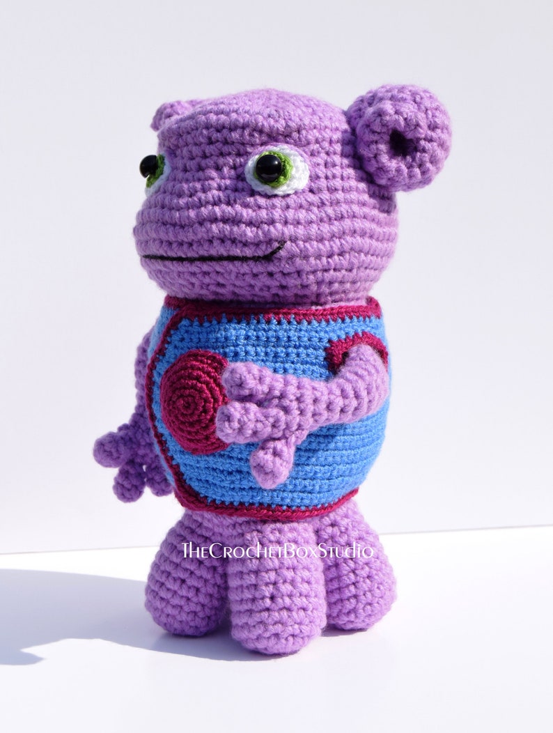 May include: Purple crocheted stuffed toy with blue and burgundy accents. The toy has large green eyes and a small smile. The toy is standing on a white surface. The text "THECROCHETBOXSTUDIO" is visible on the toy's chest.