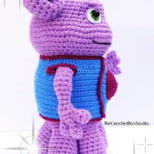 May include: A crocheted purple plush toy with a blue vest and a red heart. The toy has a large head with a green eye and a small mouth.